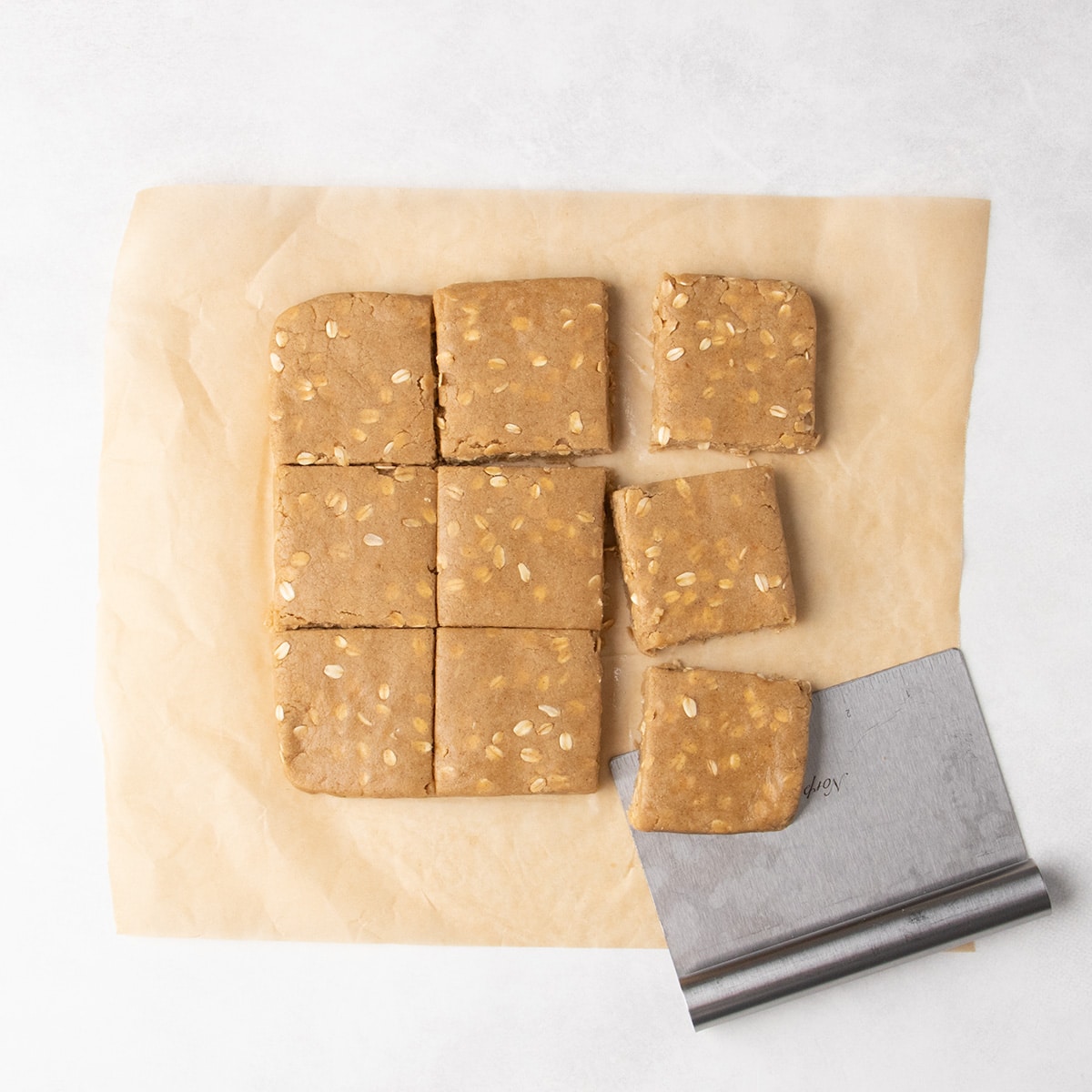 Nine raw square pieces of oat-speckled dough rest on parchment paper beside a metal bench scraper.