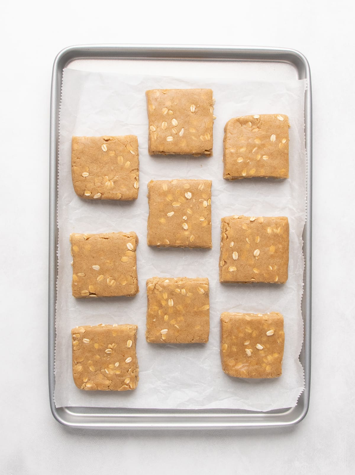 Nine raw square pieces of oat-speckled dough are spaced evenly across a parchment-lined metal baking sheet.