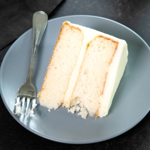 A slice of two-layer gluten free white cake with white frosting sits on a gray plate beside a fork.