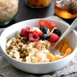 Closeup image of a white bowl that holds muesli recipe ingredients, including oats, nuts, seeds, coconut flakes, dried apricots, strawberries, raspberries, and blueberries with a spoon.