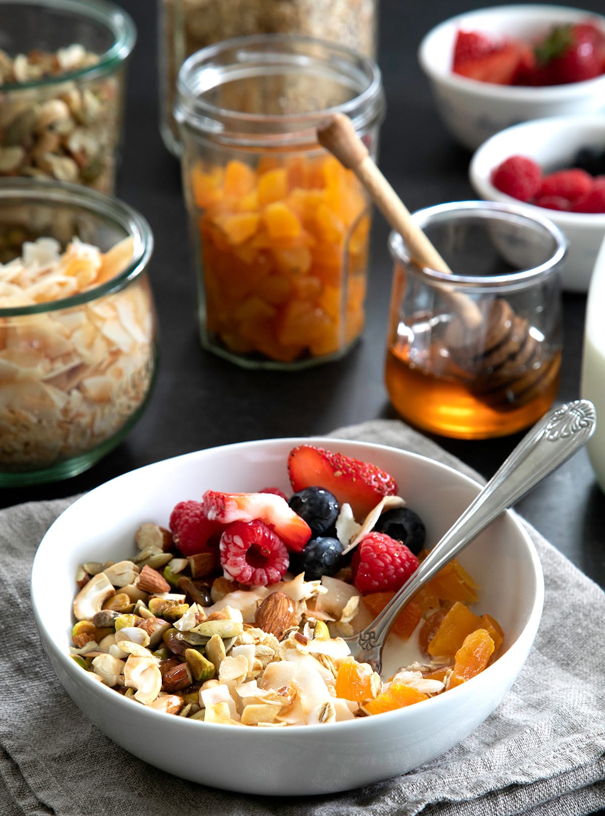 A bowl of oats, nuts, seeds, coconut flakes, dried apricots, strawberries, raspberries, and blueberries sits beside jars of the ingredients and a glass of honey.
