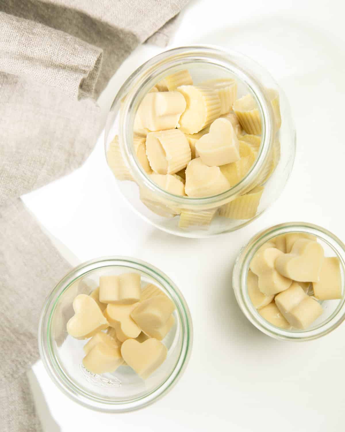 Three glass jars hold pale white chocolate pieces in heart, round, and ridged shapes on a white surface beside a beige cloth.