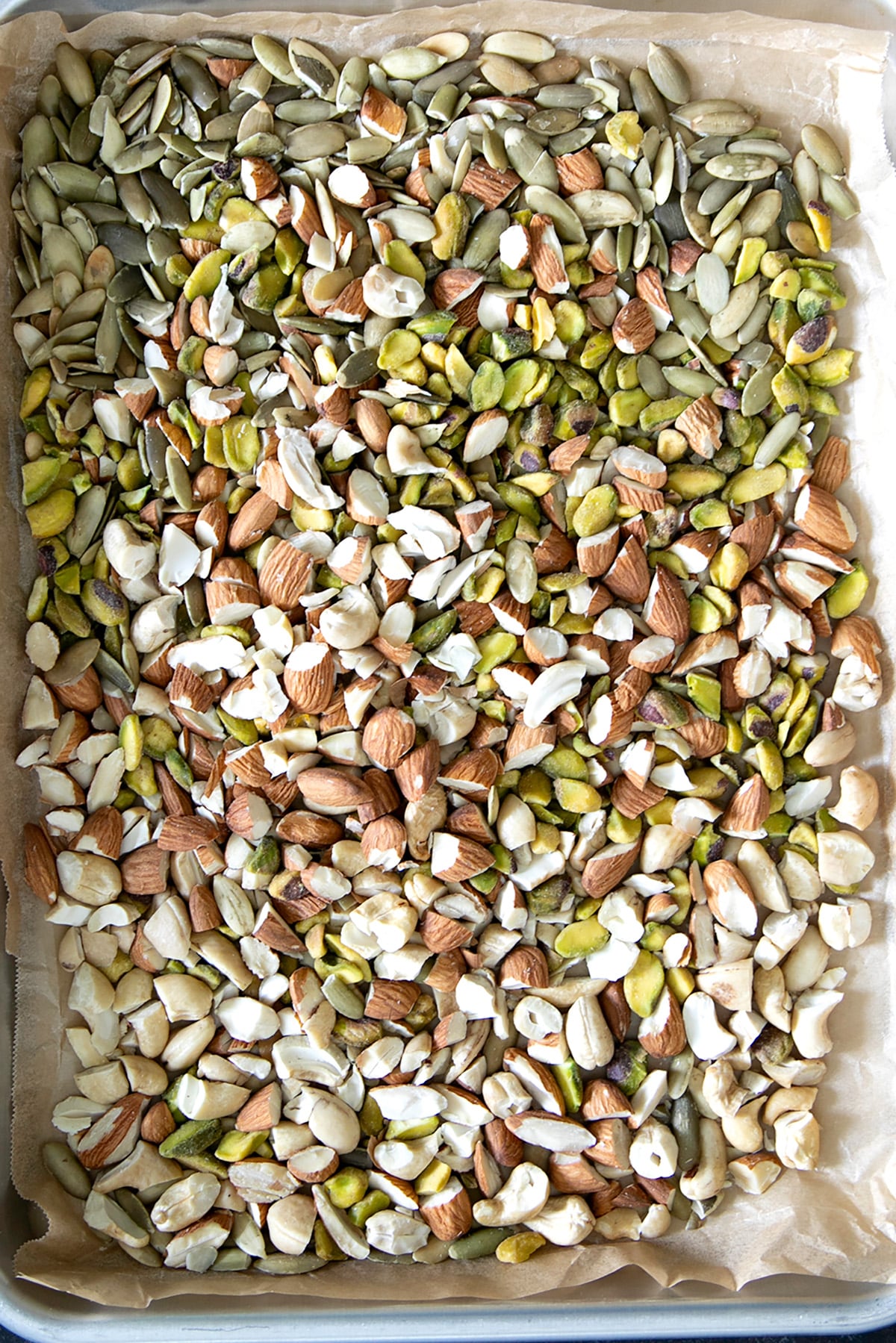Chopped almonds, cashews, pistachios, and pumpkin seeds cover a parchment-lined baking sheet.