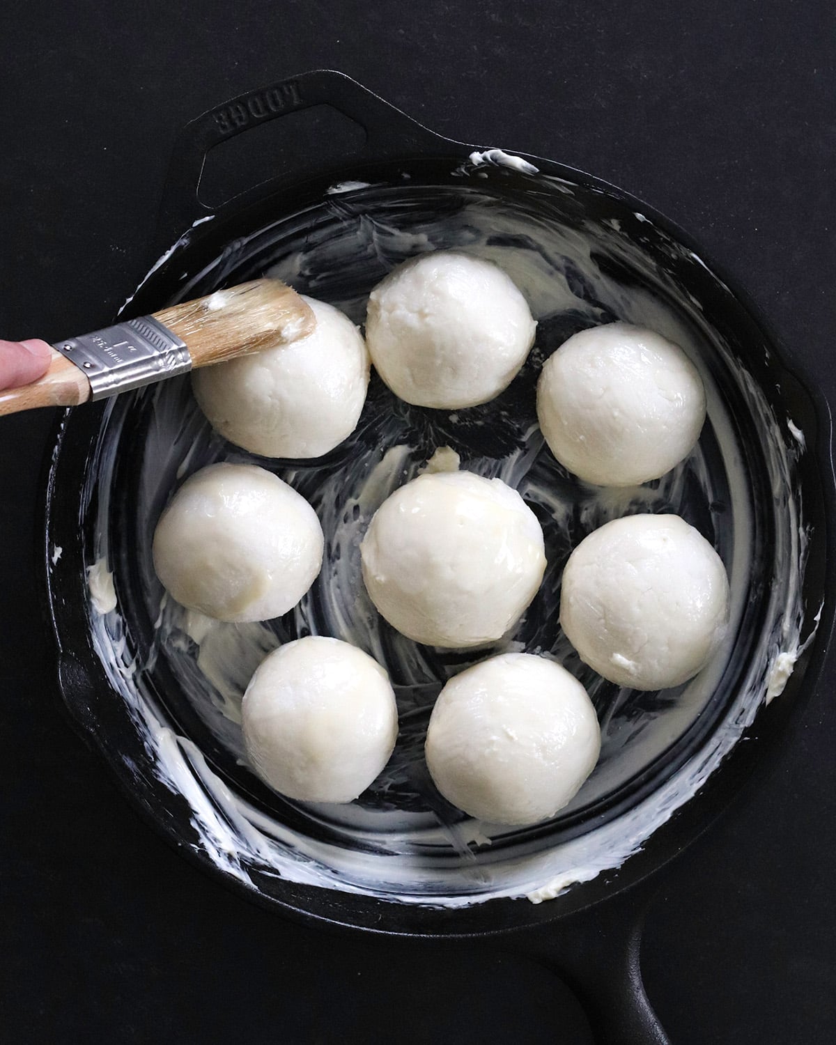 A hand uses a pastry brush to coat with softened butter one of eight round raw dough balls arranged in a greased black cast iron skillet.