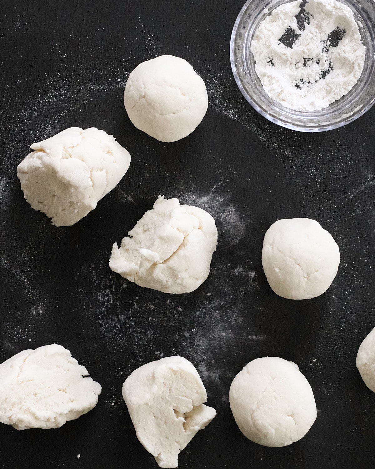 Portions of white dough sit on a dark surface beside two smooth dough balls and a bowl of white flour.