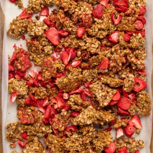 Closeup overhead image of strawberry granola clusters and freeze-dried strawberry pieces filling a parchment-lined baking sheet.