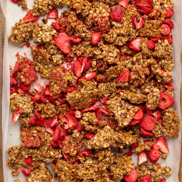 Closeup overhead image of strawberry granola clusters and freeze-dried strawberry pieces filling a parchment-lined baking sheet.