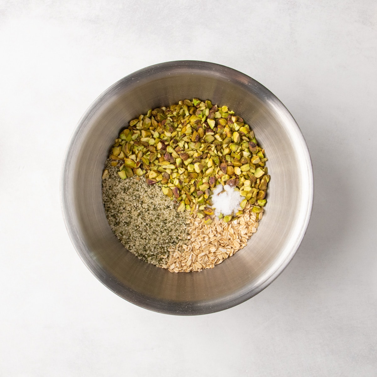 Rolled oats, chopped pistachios, small hemp seeds, and a mound of white salt granules sit separately in a large metal mixing bowl.