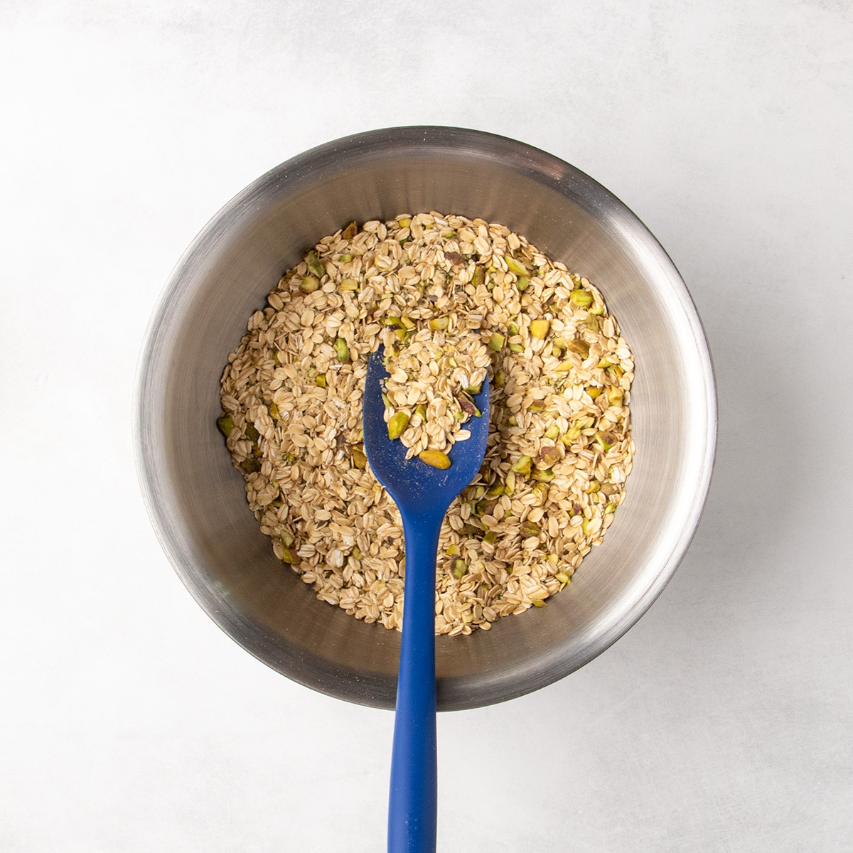 A blue spatula rests in a metal bowl after the oats, pistachios, seeds, and salt have been mixed together.