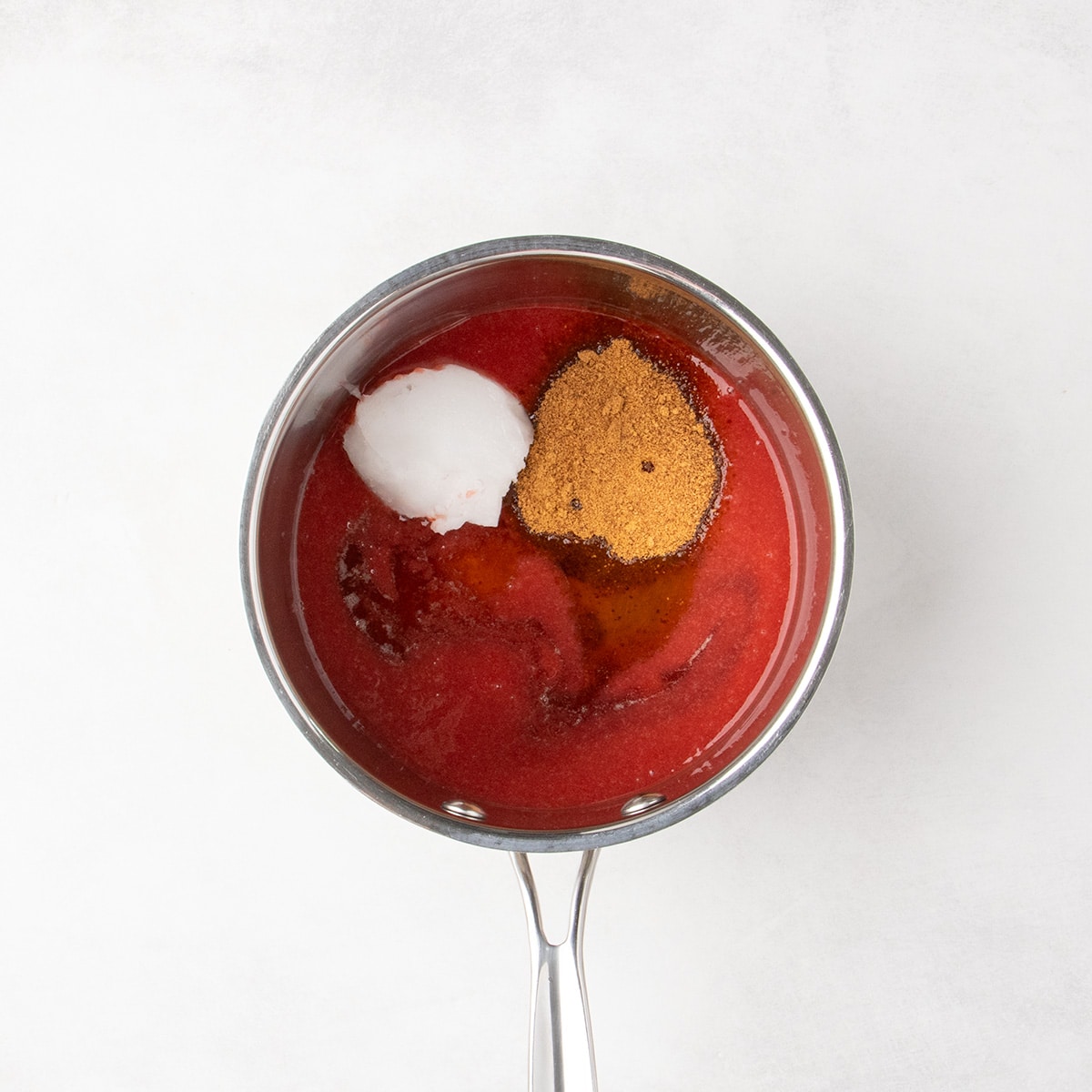 A saucepan holds a red strawberry puree liquid with white solid coconut oil, brown granules of coconut sugar, and a small pool of amber liquid on top.