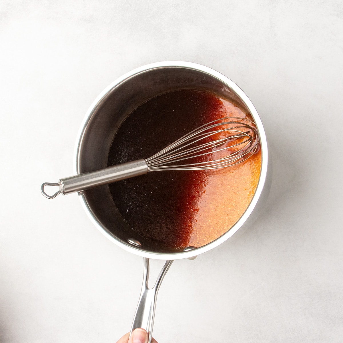 A whisk rests in a saucepan filled with a smooth dark reddish-brown liquid while the saucepan is being tipped to the side.