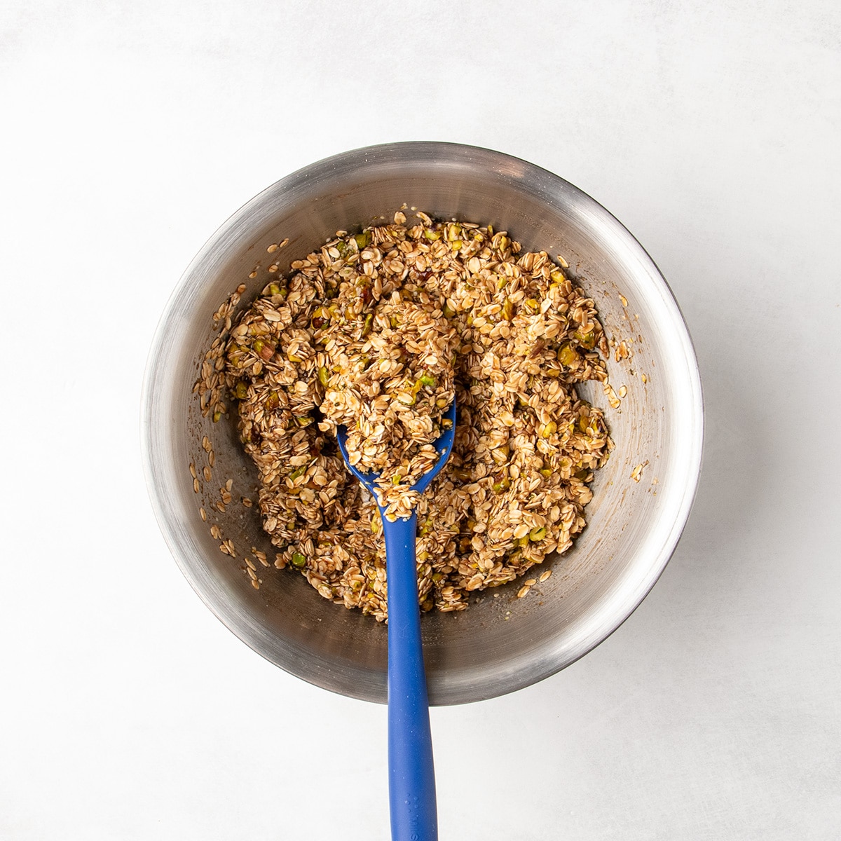A blue spatula rests in a metal bowl of oats, pistachios, and seeds coated with the dark liquid.