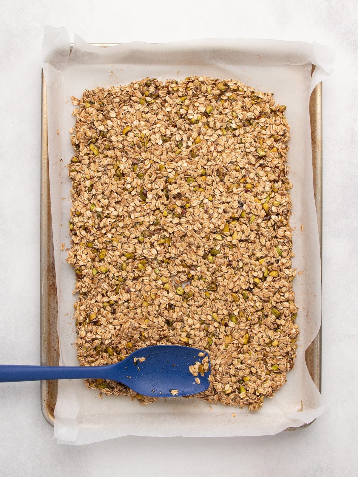 A blue spatula spreads the coated oat mixture over a parchment-lined baking sheet.