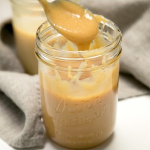 closeup image of a spoon lifting a thick tan mixture of sweetened condensed milk above a glass jar filled with the same mixture.