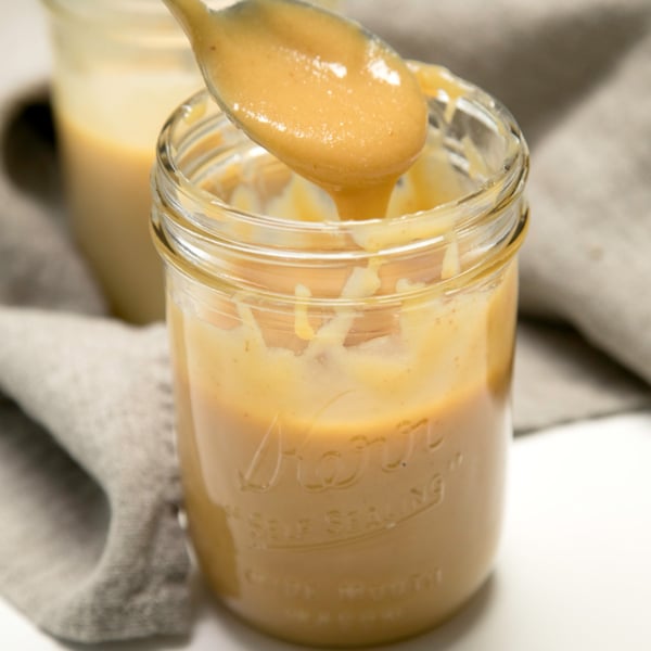 closeup image of a spoon lifting a thick tan mixture of sweetened condensed milk above a glass jar filled with the same mixture.