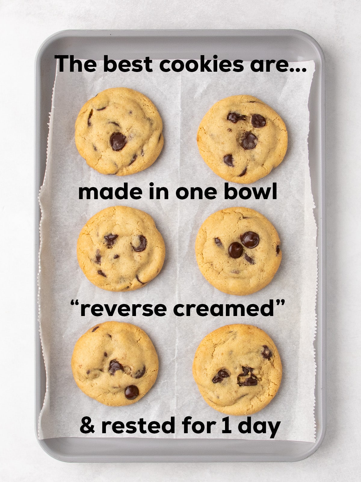 Overhead image of 6 golden brown gluten free chocolate chip cookies with slightly more browned edges on metal baking sheet with white paper lining with words "The best cookies are... "made in one bowl, reverse creamed & rested for 1 day."