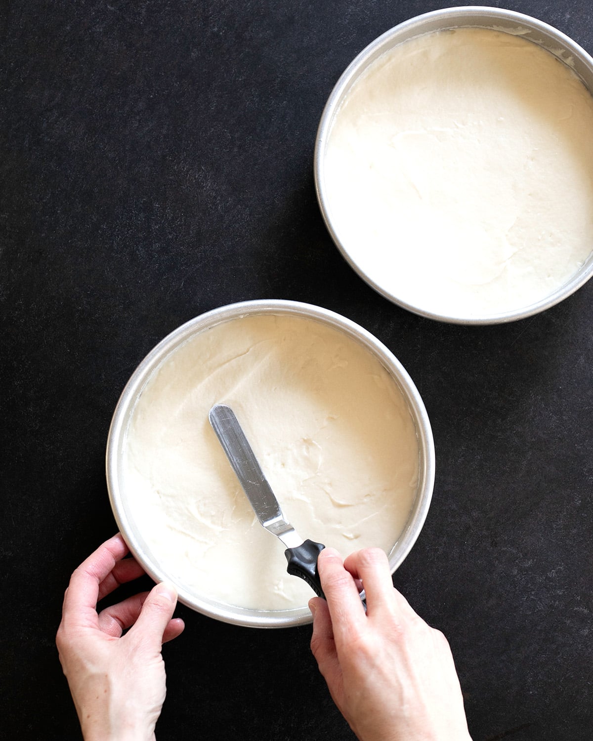Two round cake pans hold smooth, pale cake batter while a hand uses an offset spatula to level the batter in one pan.