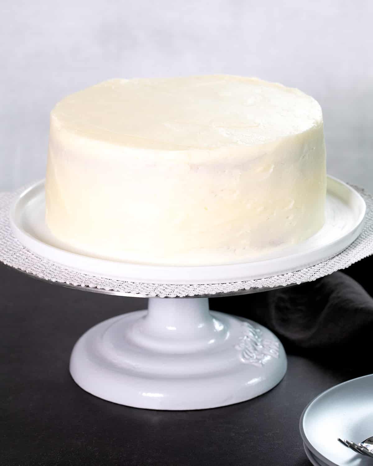 A fully frosted round white cake sits on a white pedestal cake stand.