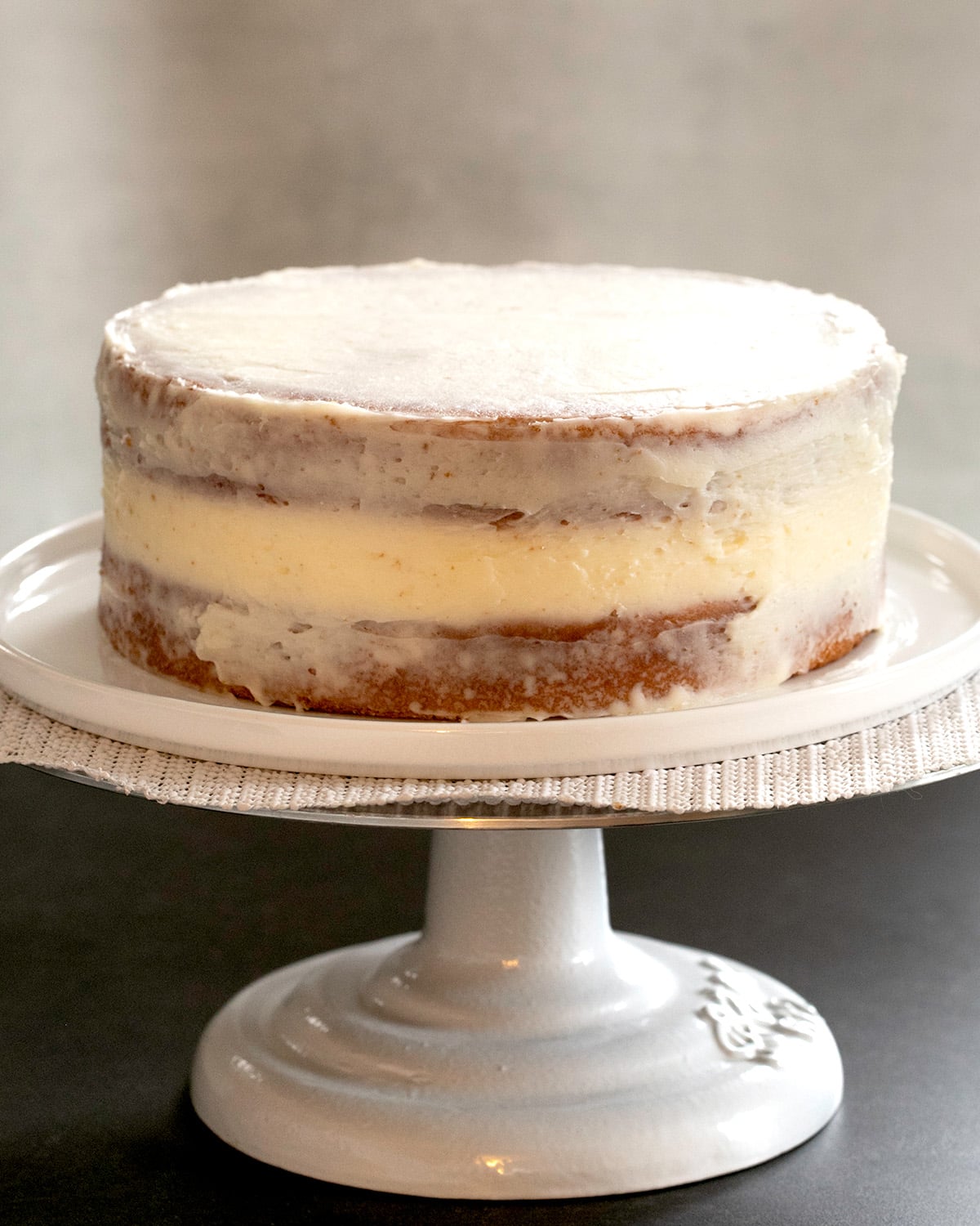 A two-layer round cake sits on a pedestal stand with a thin layer of white frosting covering the top and sides.