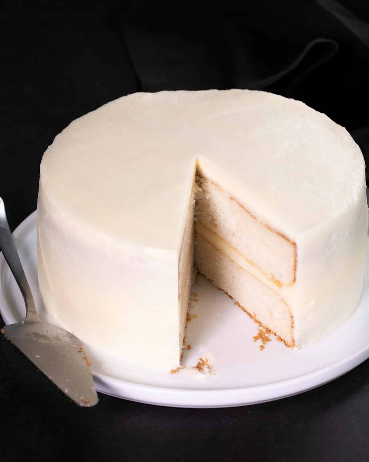 A two-layer white frosted cake sits on a white plate with a slice removed and a cake server resting beside it.