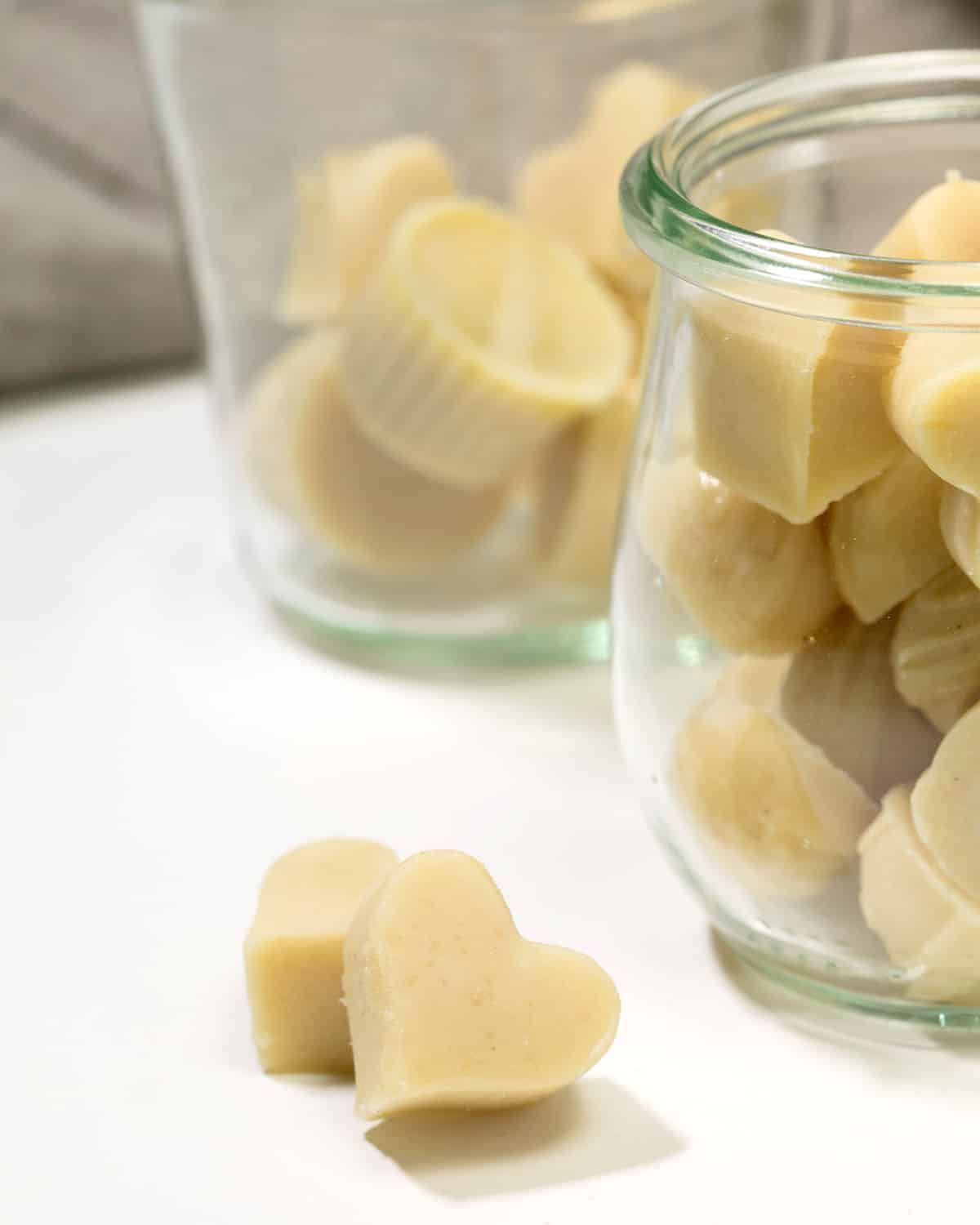 Two heart-shaped white chocolate pieces rest on a white surface beside glass jars filled with more pale white chocolate pieces.