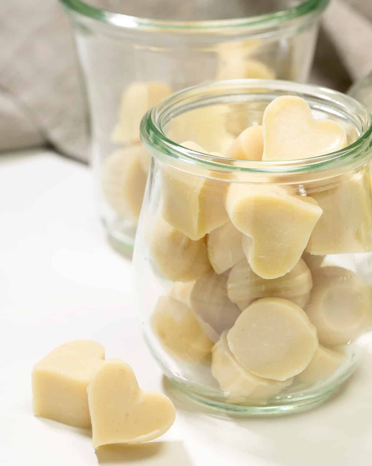A glass jar filled with pale white chocolate pieces sits in front of another jar, with two heart-shaped pieces resting on the white surface.
