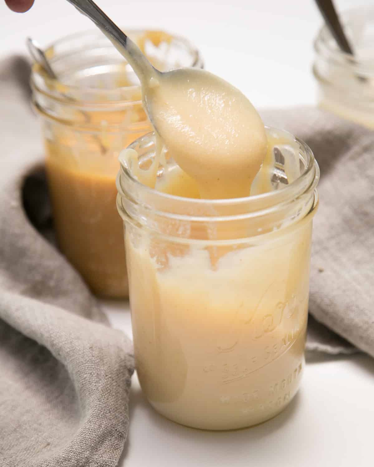 A spoon lifts a thick pale cream-colored mixture above a glass jar, with another jar blurred in the background.