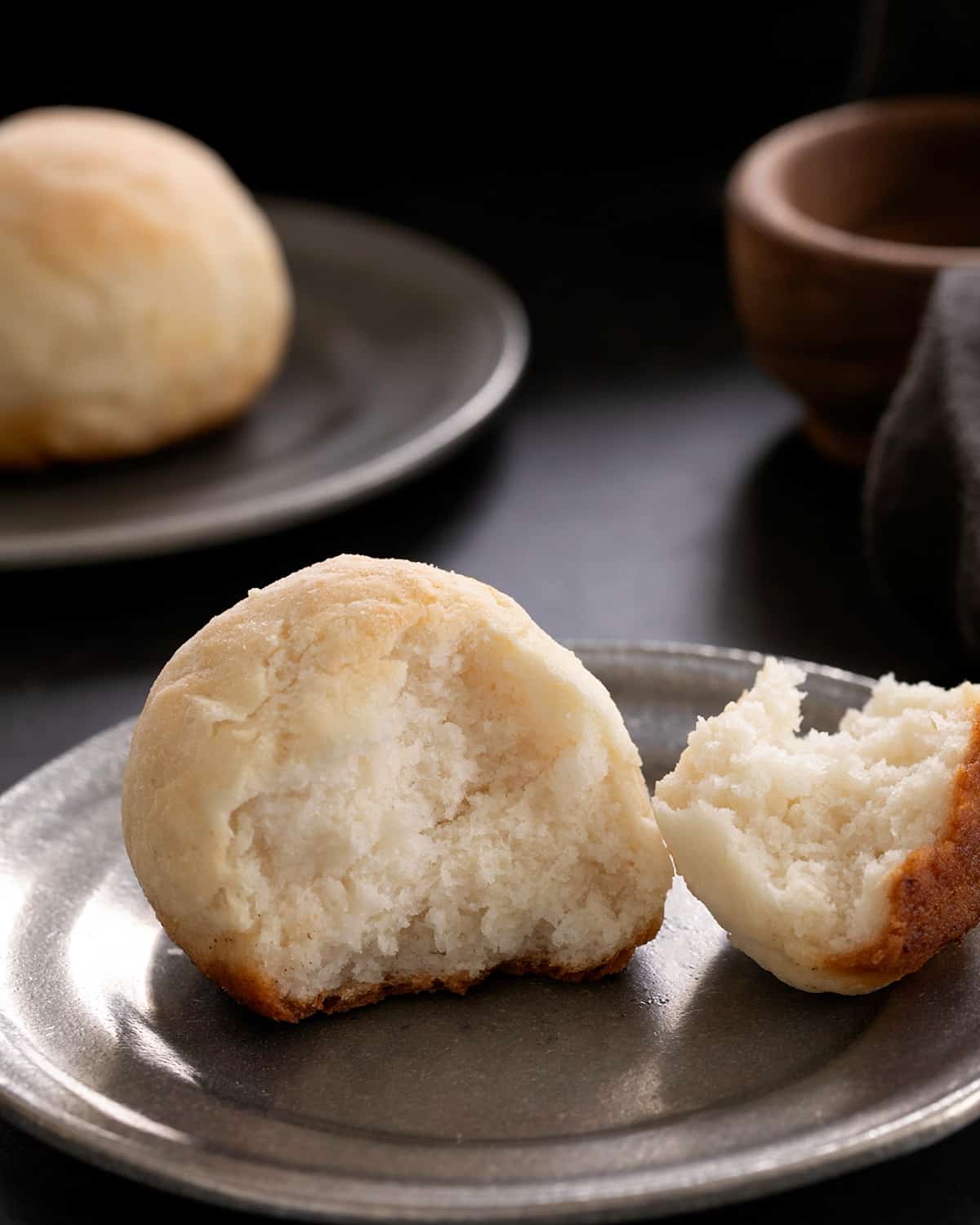 A torn-open gluten free yeast free dinner roll shows a soft white interior on a dark plate, with another roll blurred in the background.