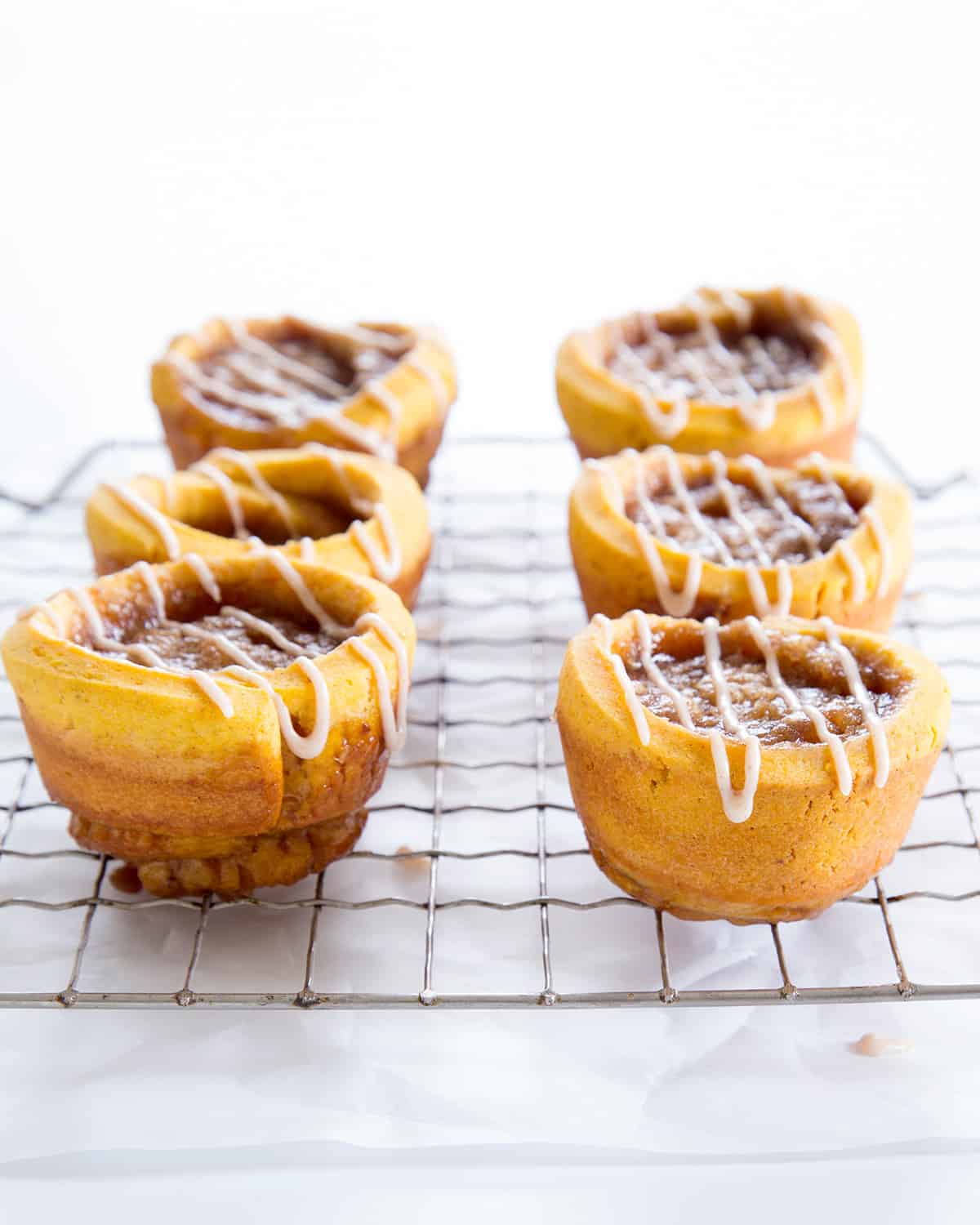 A wire rack with 6 brown-orange pumpkin cinnamon rolls with white icing drizzled on top on white paper