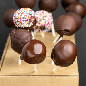 closeup image of gluten free cake pops with chocolate coating stuck into brown cardboard box as they dry, some with colorful nonpareils
