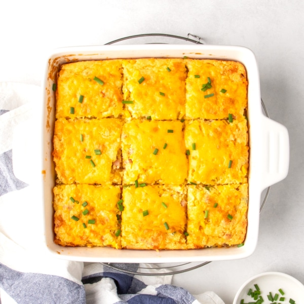 Closeup overhead image of gluten free corn casserole cut into 9 equal squares and topped with melted cheddar cheese and chopped chives in a white square casserole dish on a round wire rack.