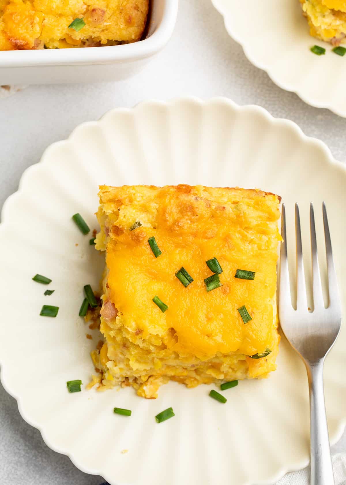 A square serving of gluten free corn casserole with melted cheddar and chopped chives sits on a white plate beside a fork.