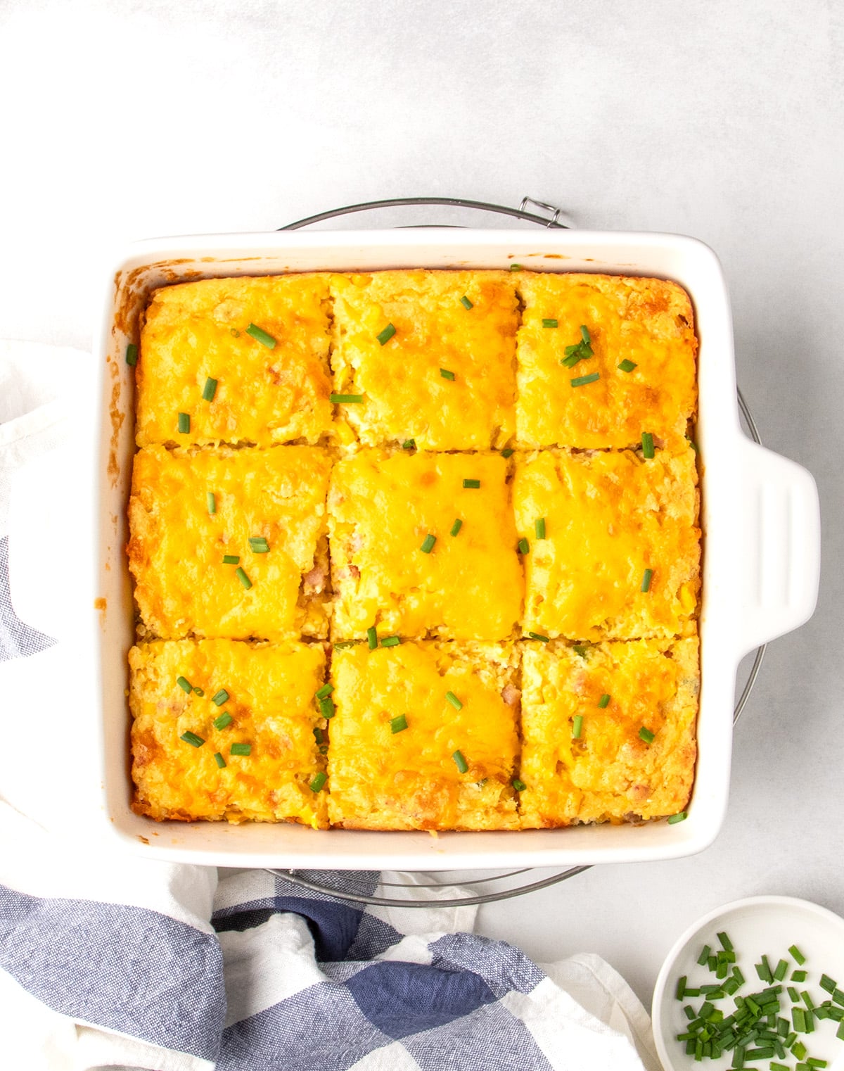 Gluten free corn casserole is cut into squares in a white square porcelain baking dish topped with melted cheddar cheese and chopped chives on a round wire rack with a blue and white cloth.