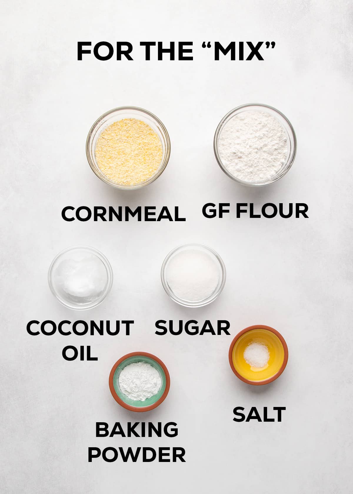 Yellow cornmeal, gluten free flour, coconut oil, sugar, baking powder, and salt are arranged and labeled on a white surface.