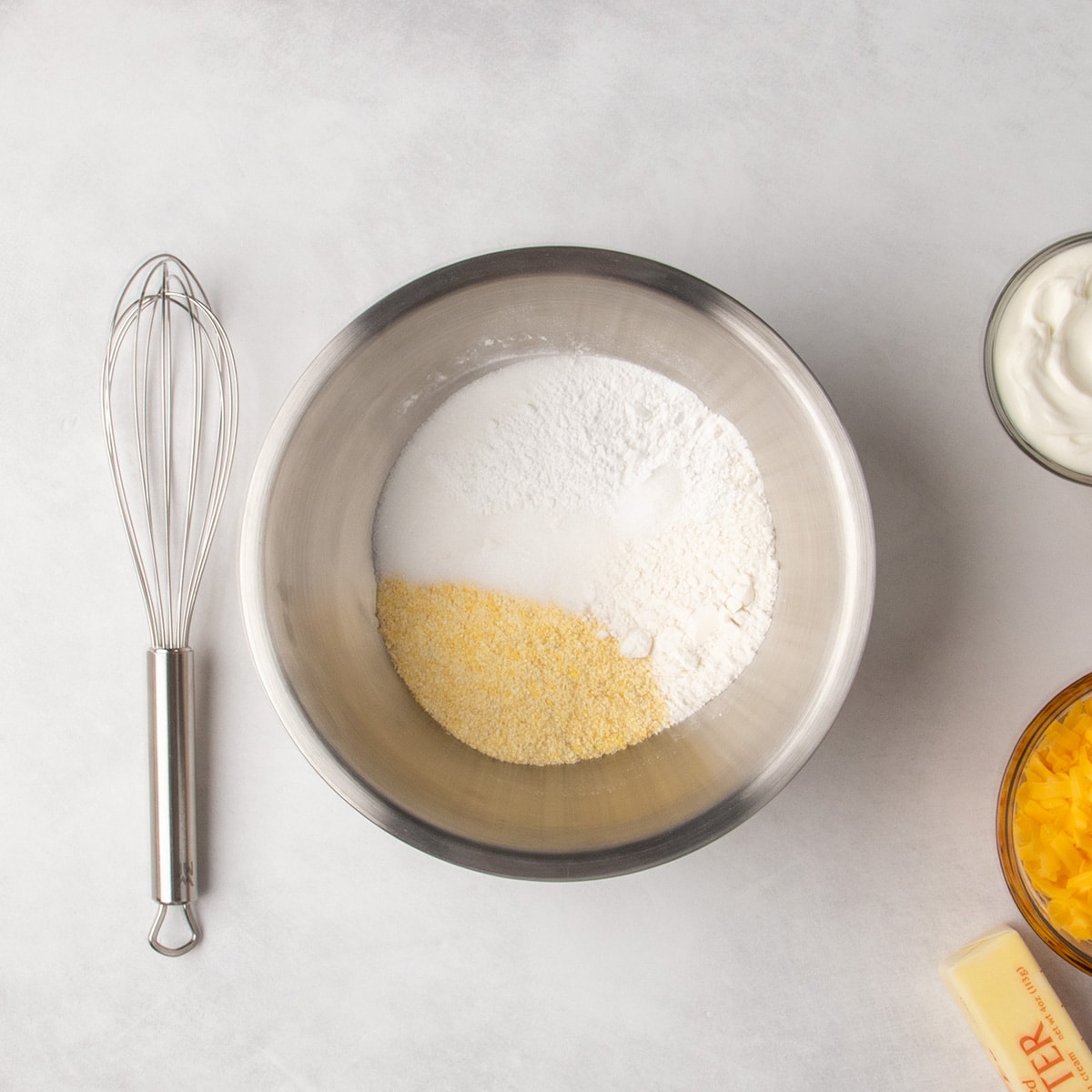 White and yellow dry ingredients sit in a metal mixing bowl with a whisk beside it.