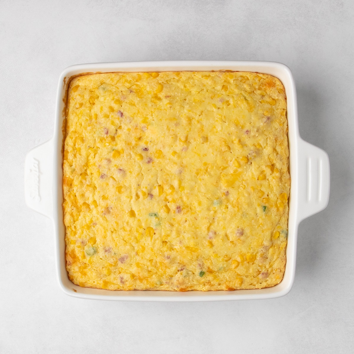 Baked corn casserole fills a square white baking dish with lightly browned edges.