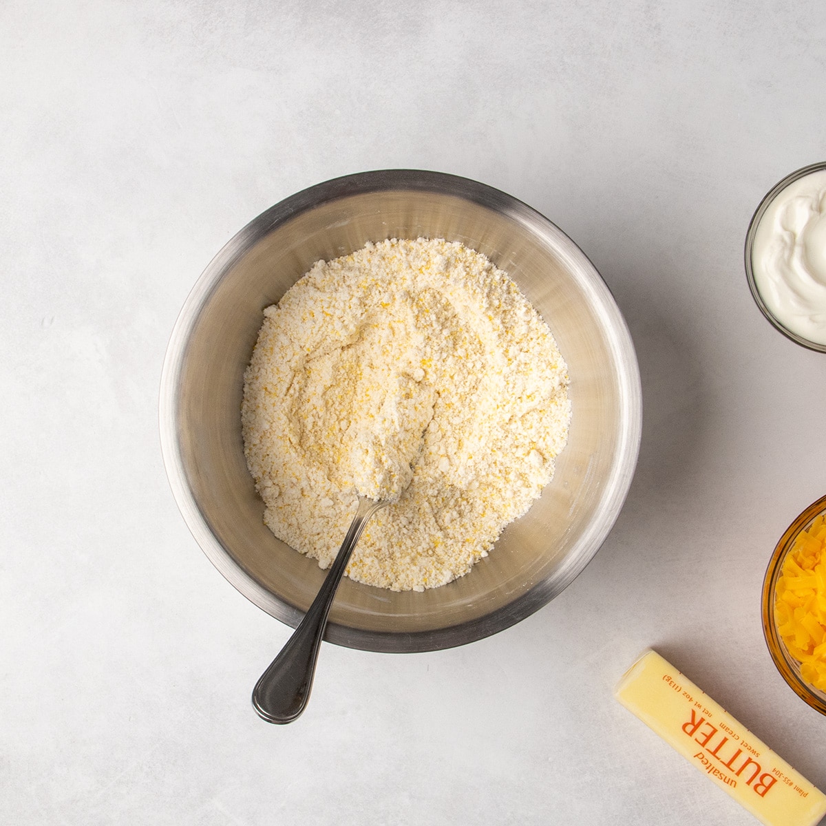 A metal fork rests in a bowl of combined dry ingredients mixed with solid coconut oil,with a stick of butter, a bowl of shredded cheese, and a bowl of sour cream nearby.