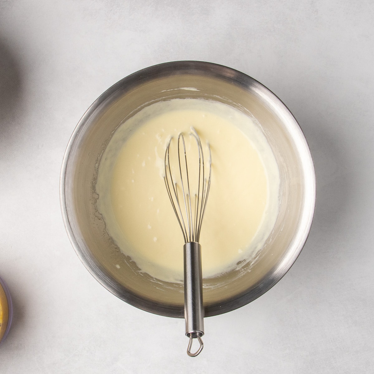 A whisk rests in a metal bowl of smooth, pale yellow wet ingredients.