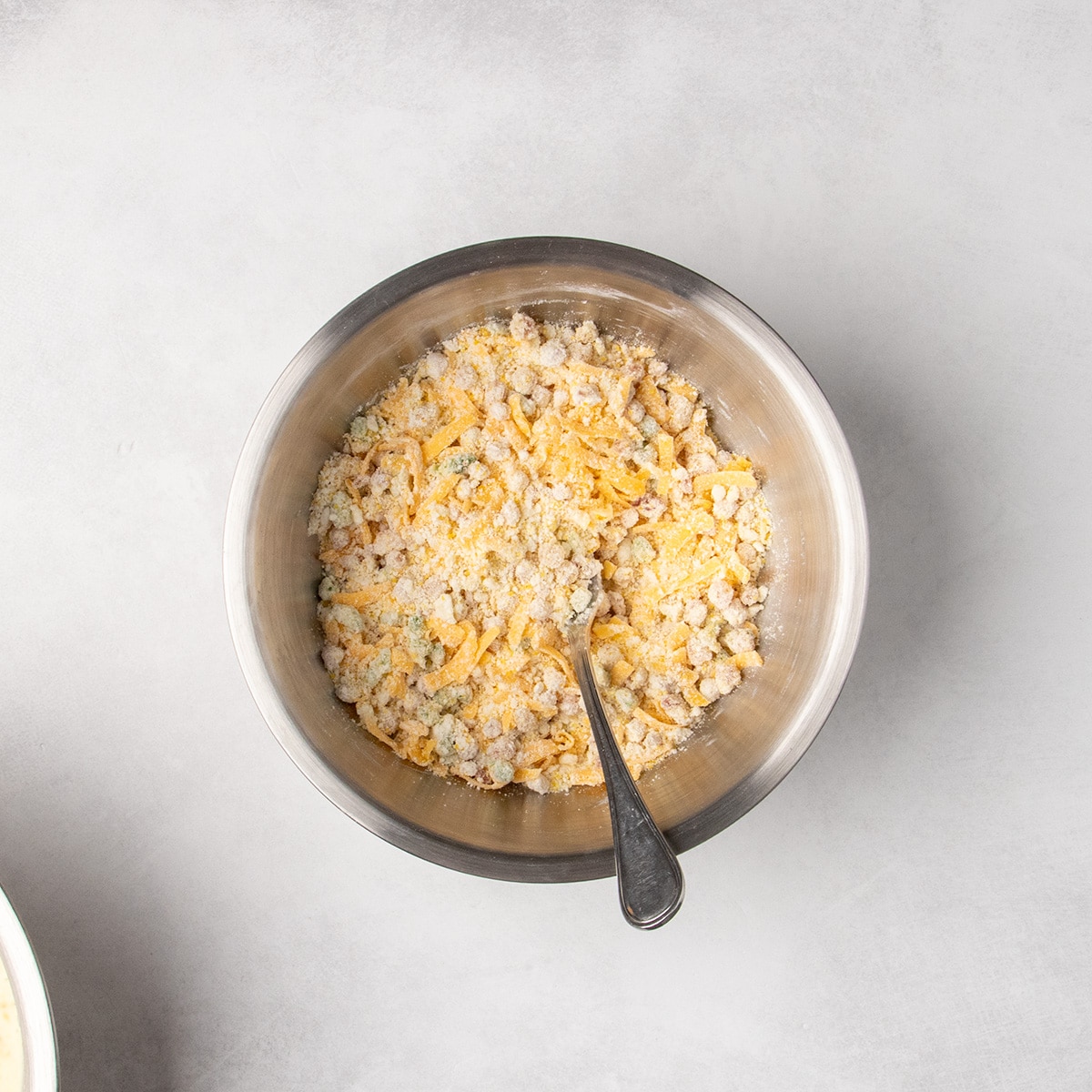 Shredded cheese, cooked pancetta and green jalapeño pepper, and dry ingredients are combined in a metal mixing bowl with a metal fork sticking out.
