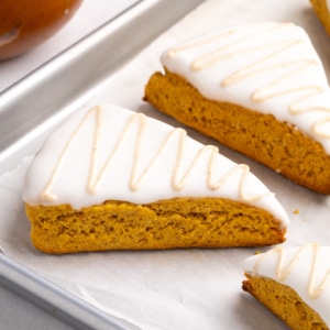 Closeup image of 3 triangular gluten free pumpkin scones with white icing and a light brown icing drizzle sit on a parchment-lined baking sheet.