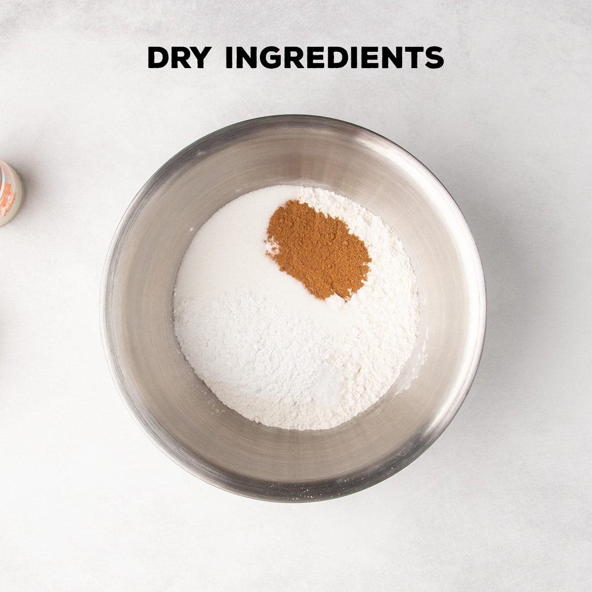 A metal mixing bowl holds flour, sugar, brown dry spice blend, and other white and light dry ingredients beneath the words “DRY INGREDIENTS.”