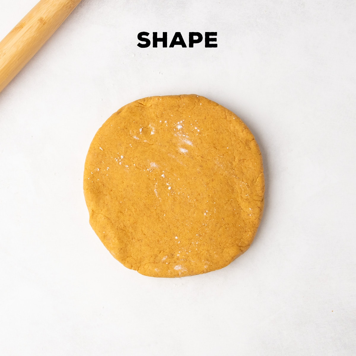 A round flat disk of orange dough sits on a white work surface beneath the word “SHAPE,” with part of a wooden rolling pin visible at the top left.