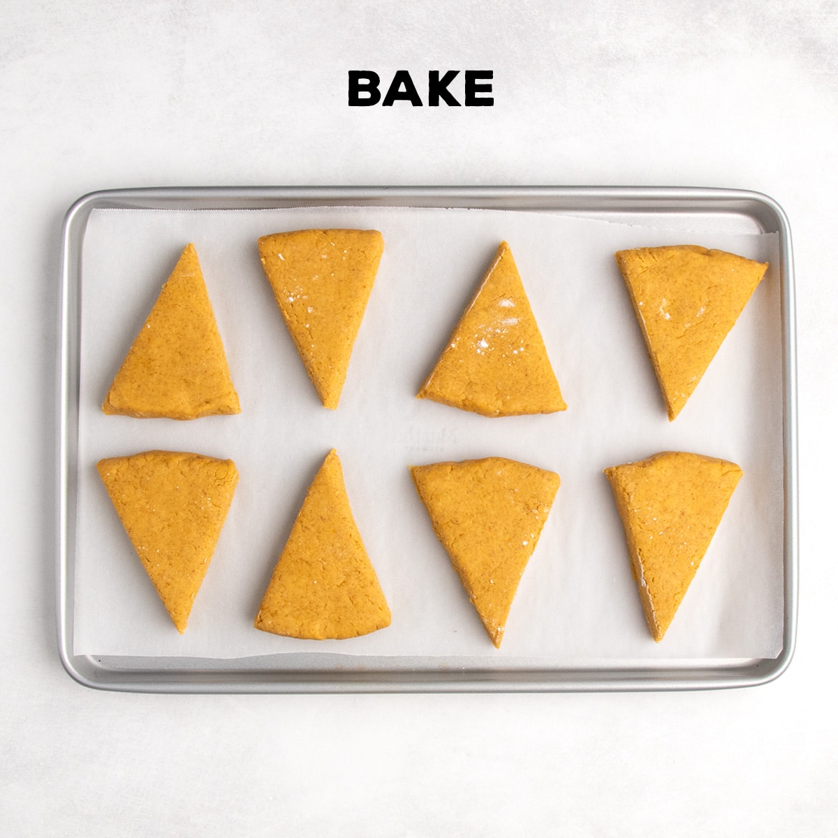 Eight triangular pieces of raw orange dough sit spaced apart on a parchment-lined baking sheet beneath the word “BAKE.”