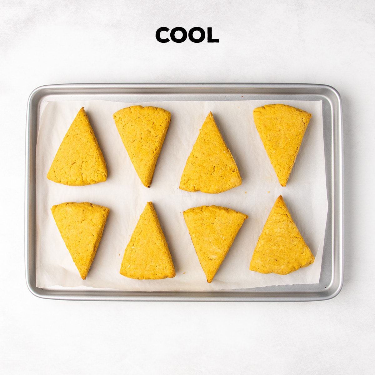 Eight baked triangular scones cool on a parchment-lined baking sheet beneath the word “COOL.”