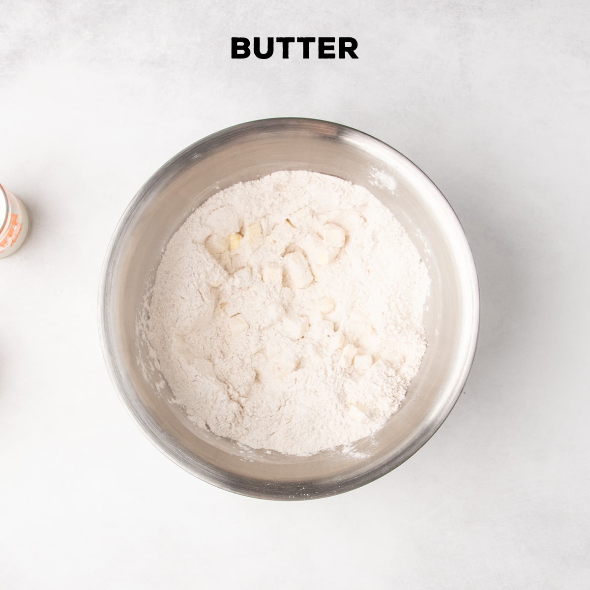 Small pieces of butter sit throughout the dry ingredients in a metal mixing bowl beneath the word “BUTTER.”