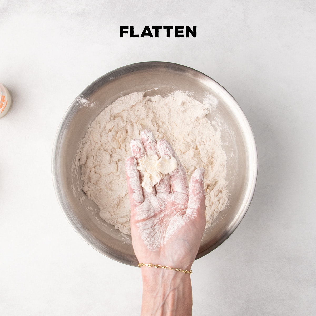 A flour-covered hand holds a flattened piece of butter over a metal bowl of dry ingredients beneath the word “FLATTEN.”