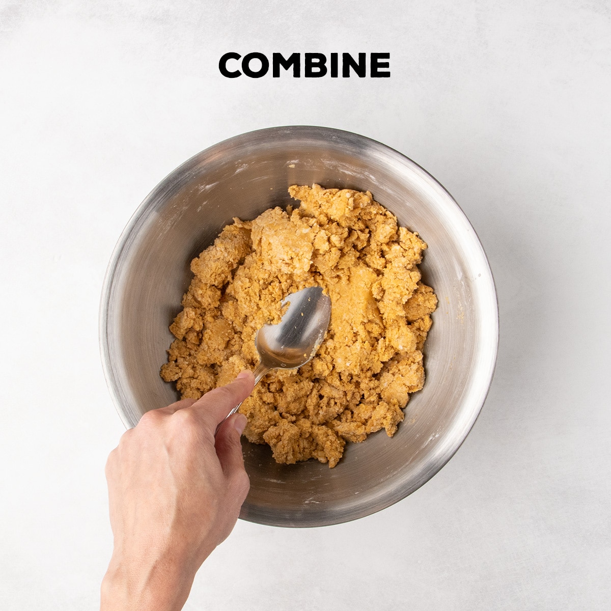 A hand uses a spoon to combine orange dough in a metal mixing bowl beneath the word “COMBINE.”