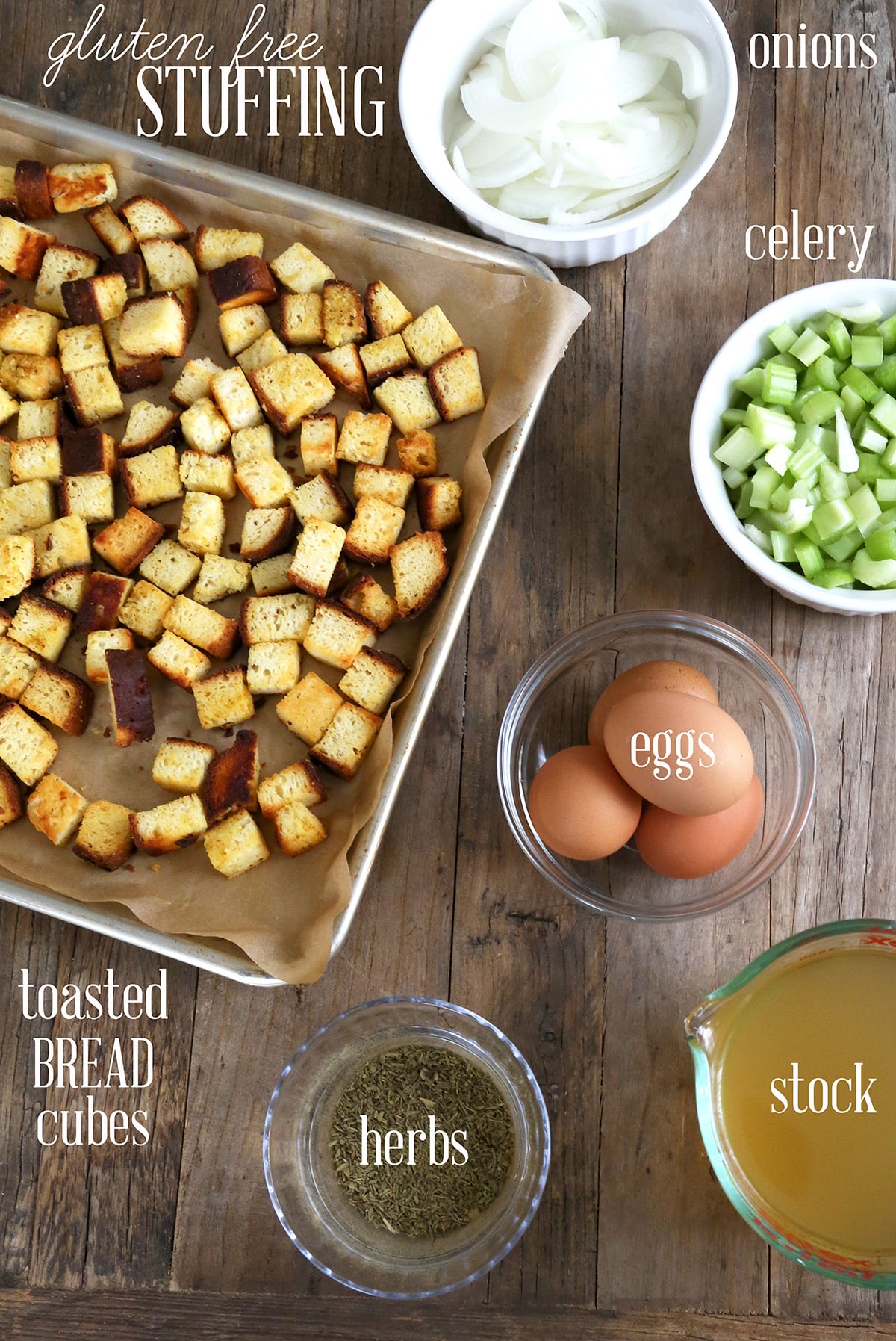 overhead image of stuffing ingredients, including dried herbs, chicken stock, gluten free bread cubes, eggs, celery and onions in small bowls and other containers on dark brown wooden board