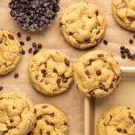Closeup image of several peanut butter protein cookies with mini chocolate chips sitting on a small light brown tray and more on brown kraft paper beside a small dish of chocolate chips, with more chips scattered around.