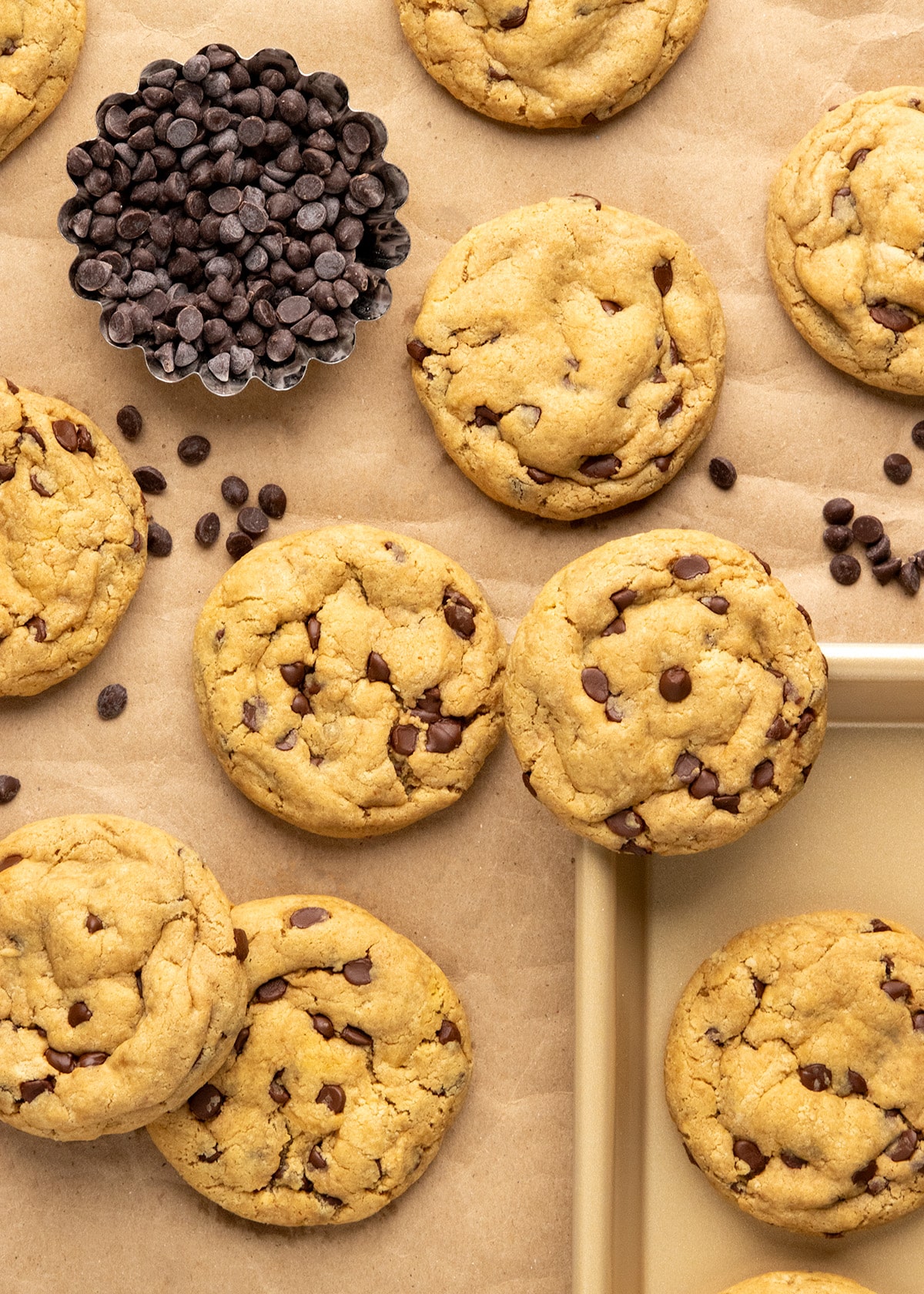 Peanut butter chocolate chip cookies sit on parchment paper around a small metal fluted dish filled with chocolate chips, with more chips scattered between them.